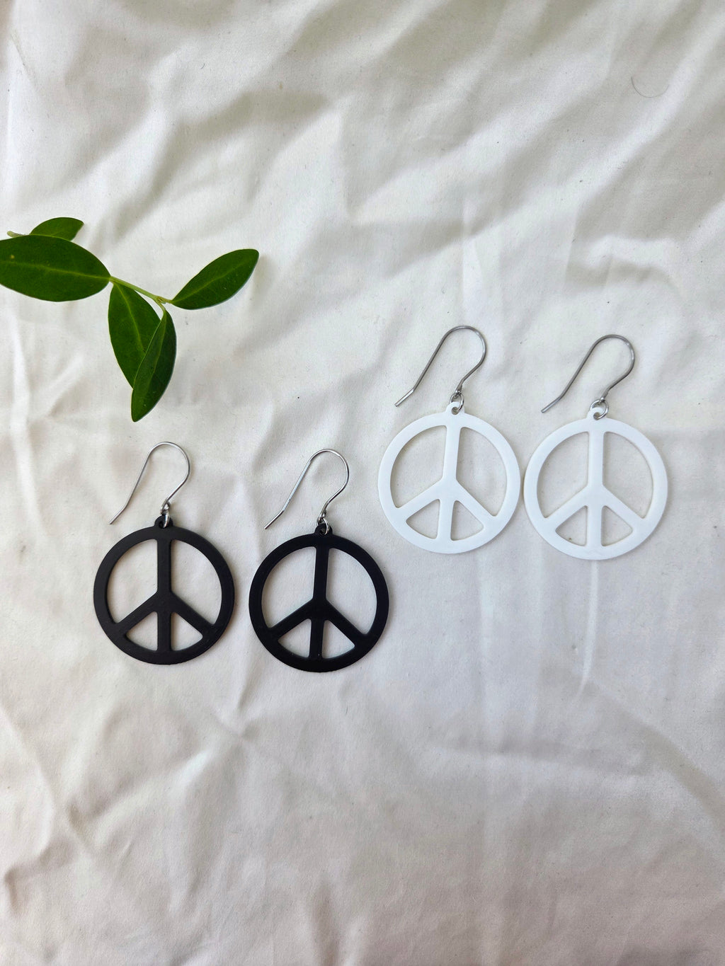 Peace sign earrings on a white fabric background with a green leaf.