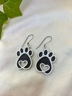 Pair of black paw print earrings on a light background with green leaves.