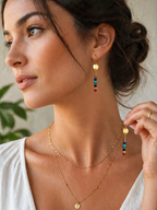 Woman wearing a gold necklace and colorful earrings, with a neutral background.