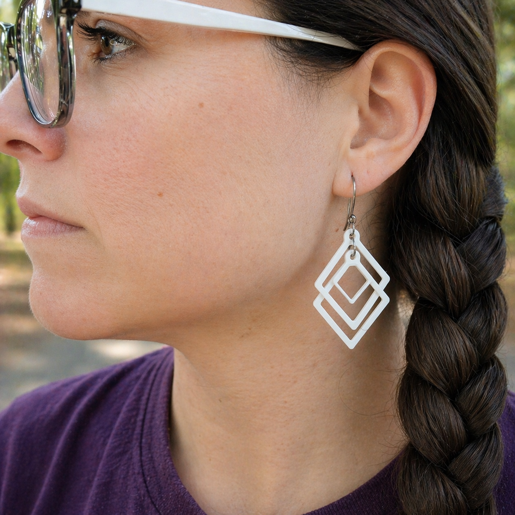 Close-up of woman wearing white sculpted open frame geometric diamond dangle earrings with braided hair and sunglasses outdoors