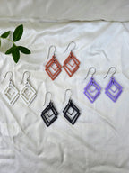 Four pairs of sculpted open frame geometric diamond earrings in white, black, terracotta, and lavender flat lay on white fabric