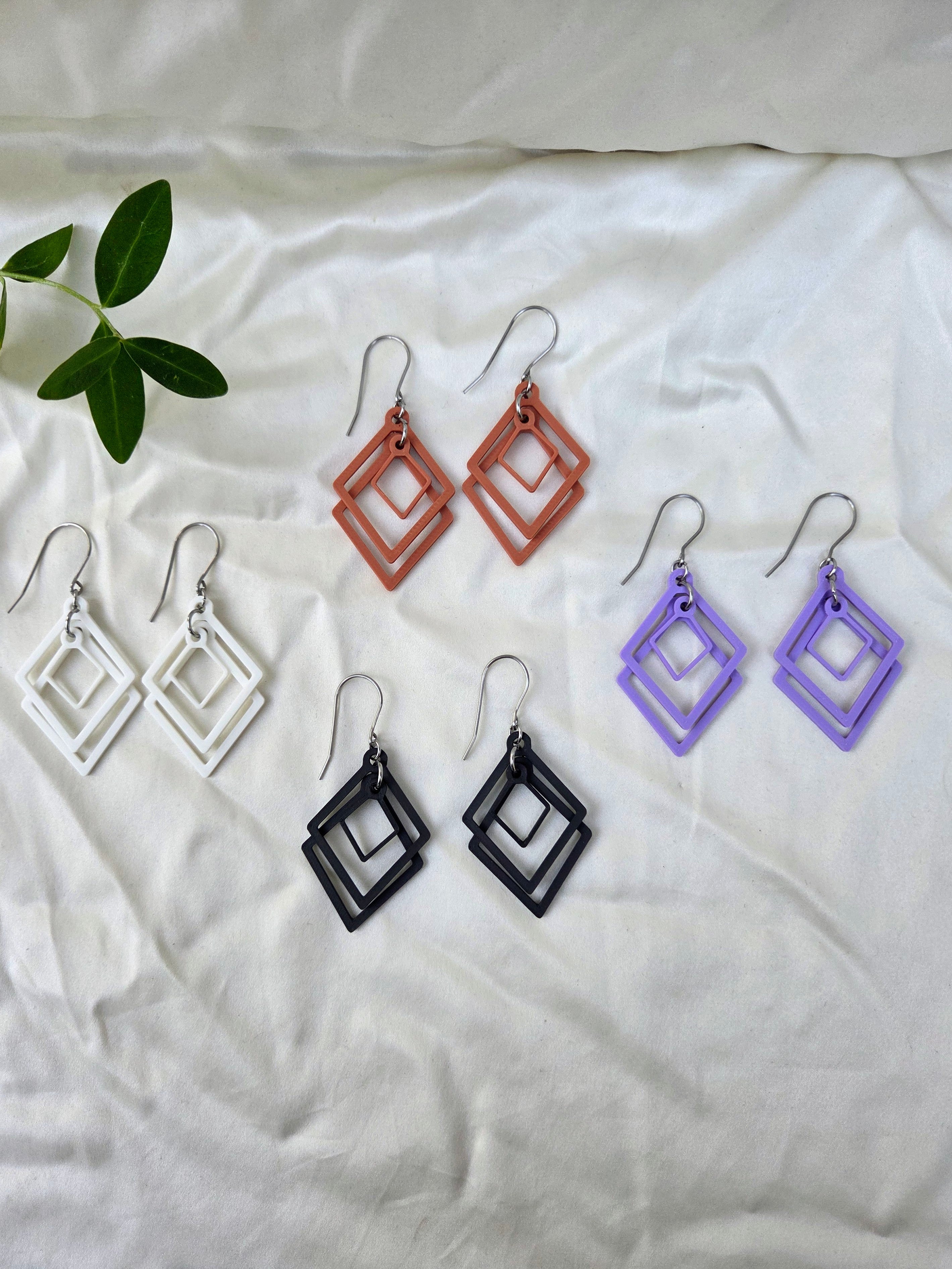 Four pairs of sculpted open frame geometric diamond earrings in white, black, terracotta, and lavender flat lay on white fabric