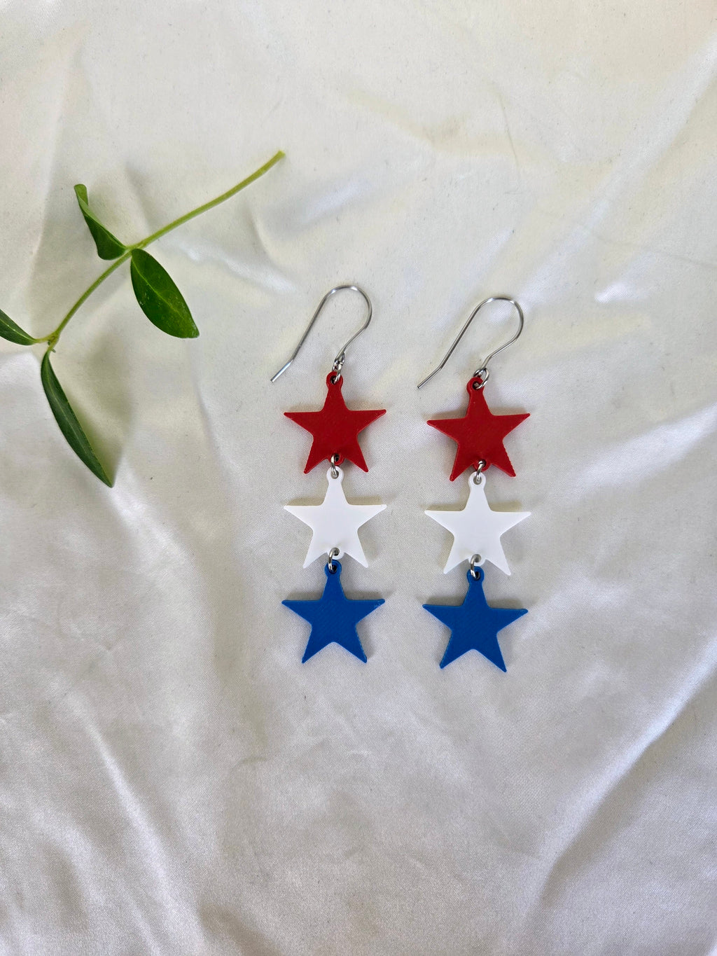 Pair of red white and blue sculpted star trio earrings flat lay on white fabric with green sprig, patriotic 4th of July jewelry