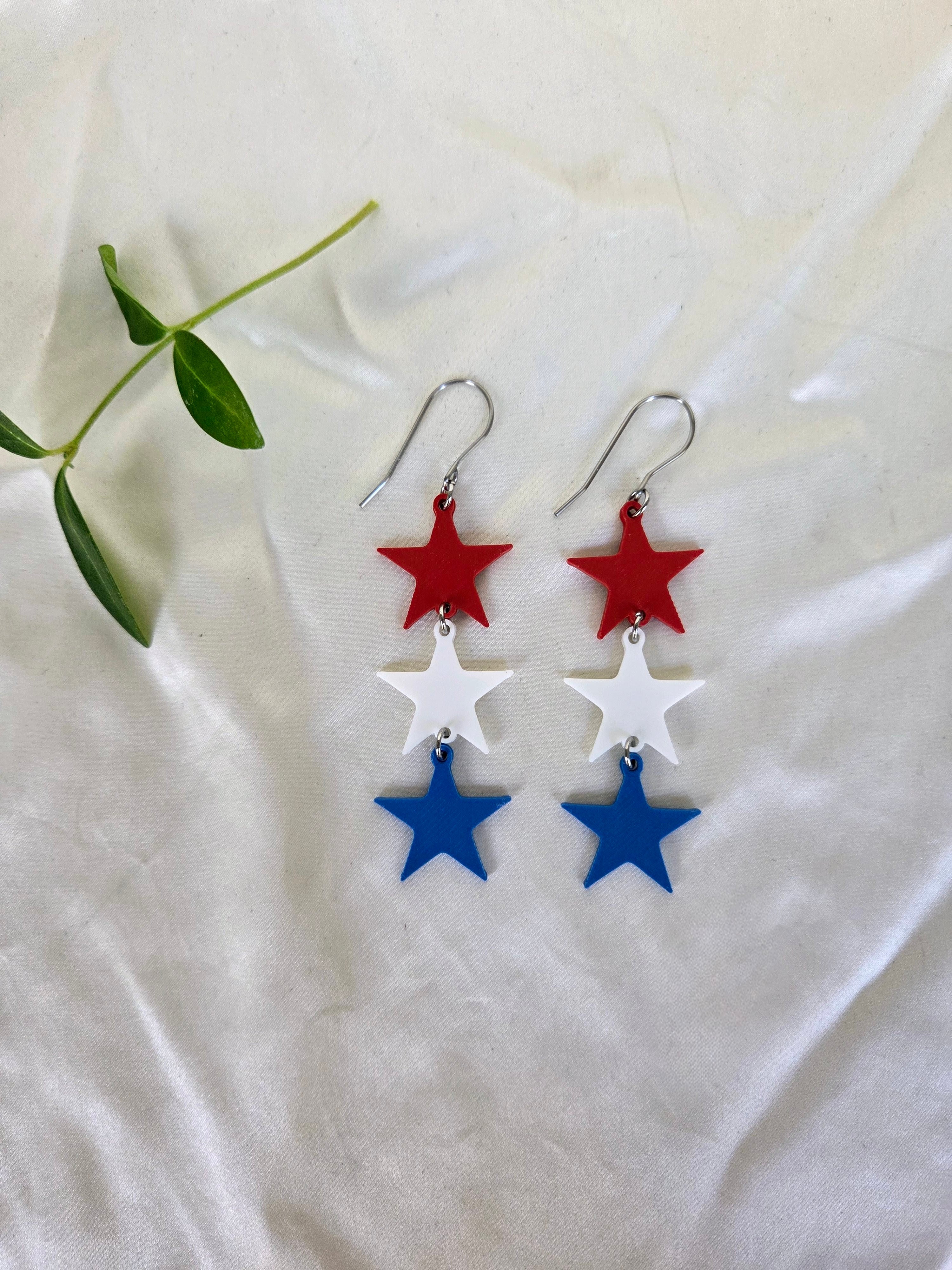 Pair of red white and blue sculpted star trio earrings flat lay on white fabric with green sprig, patriotic 4th of July jewelry