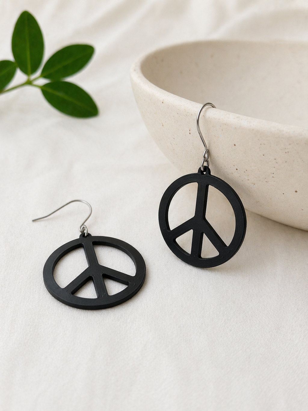 Black peace symbol earrings on a light surface with a bowl and green leaves in the background.