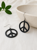 Black peace symbol earrings on a light surface with a bowl and green leaves in the background.