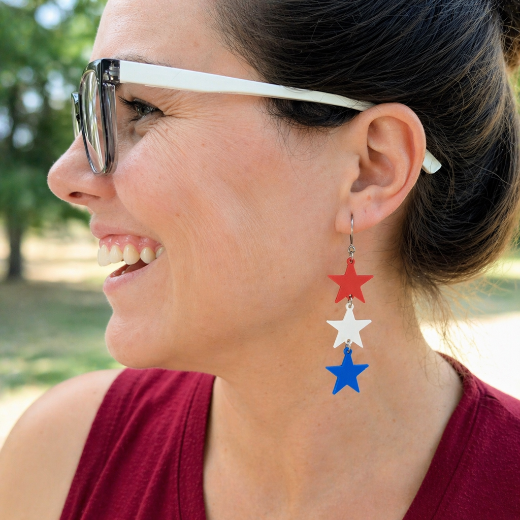 Woman wearing red white and blue sculpted star trio dangle earrings outdoors, patriotic cascading star jewelry with silver French hook