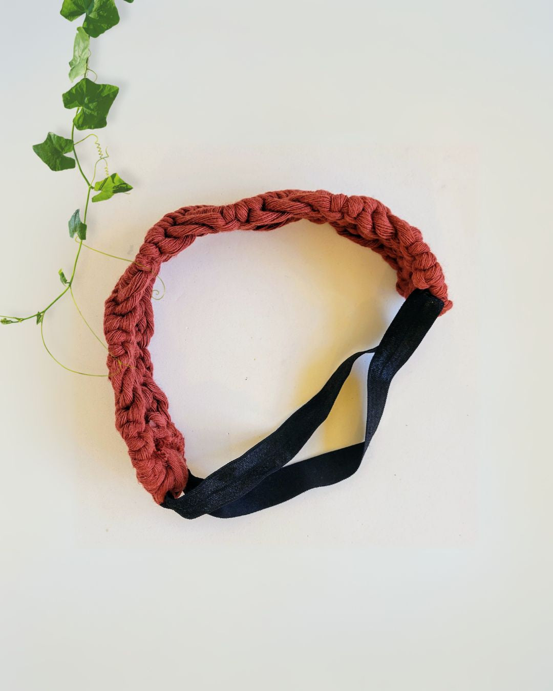 Rust red macramé braided headband with black elastic back, flat lay on white background with ivy leaves