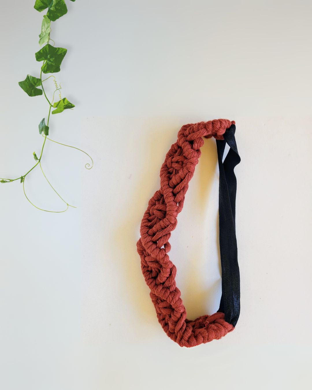 Rust red macramé braided headband with black elastic back, flat lay on white background with ivy leaves