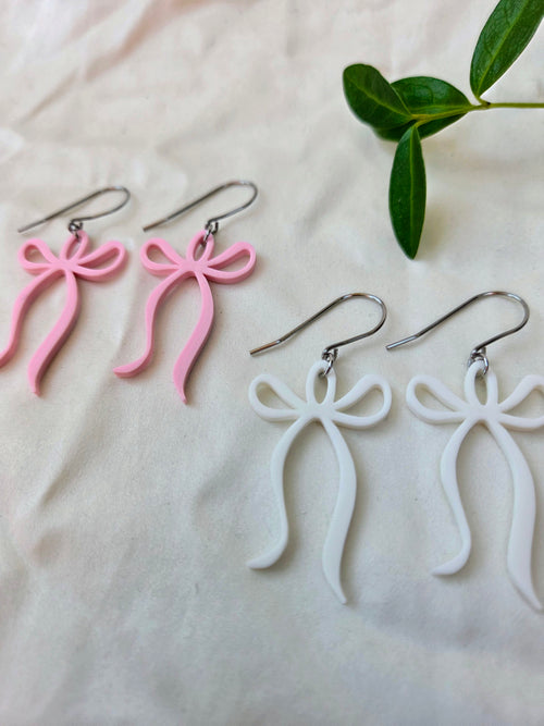 Two pairs of sculpted bow dangle earrings in blush pink and white with flowing ribbon tails, silver French hook, flat lay on white fabric with green leaves