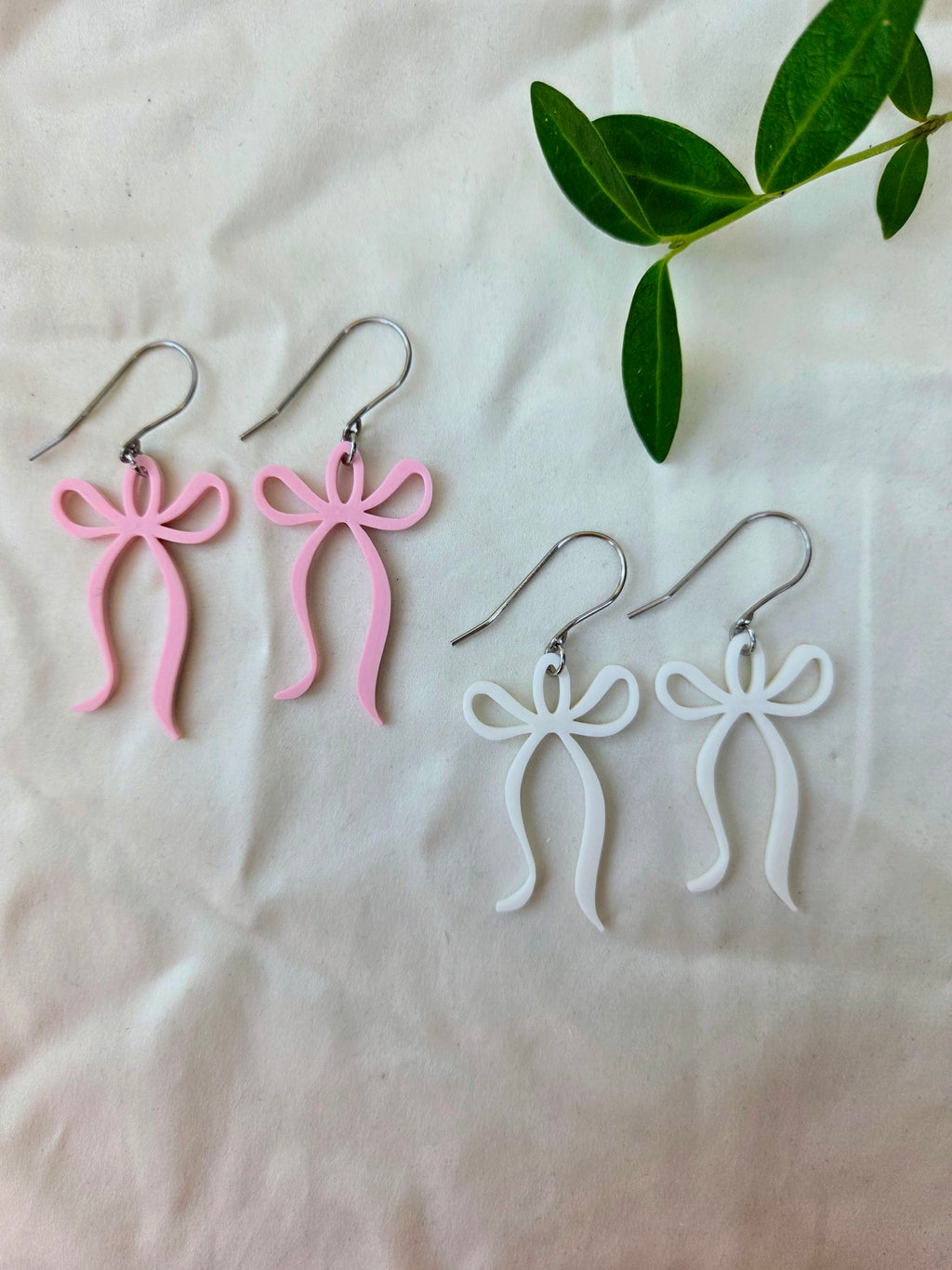 Pink and white bow-shaped earrings on a light fabric background with green leaves.