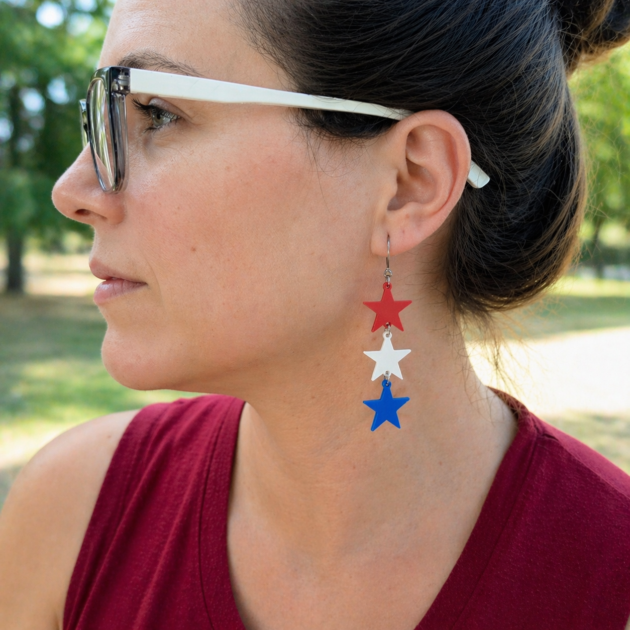 Woman wearing red white and blue sculpted star trio dangle earrings outdoors, patriotic cascading star jewelry with silver French hook